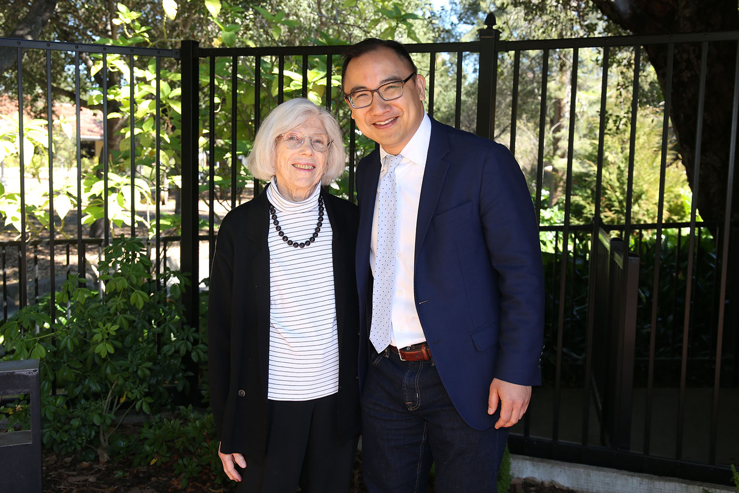 Miriam Roland and Daniel Ho posing outdoors by a black metal fence and greenery.