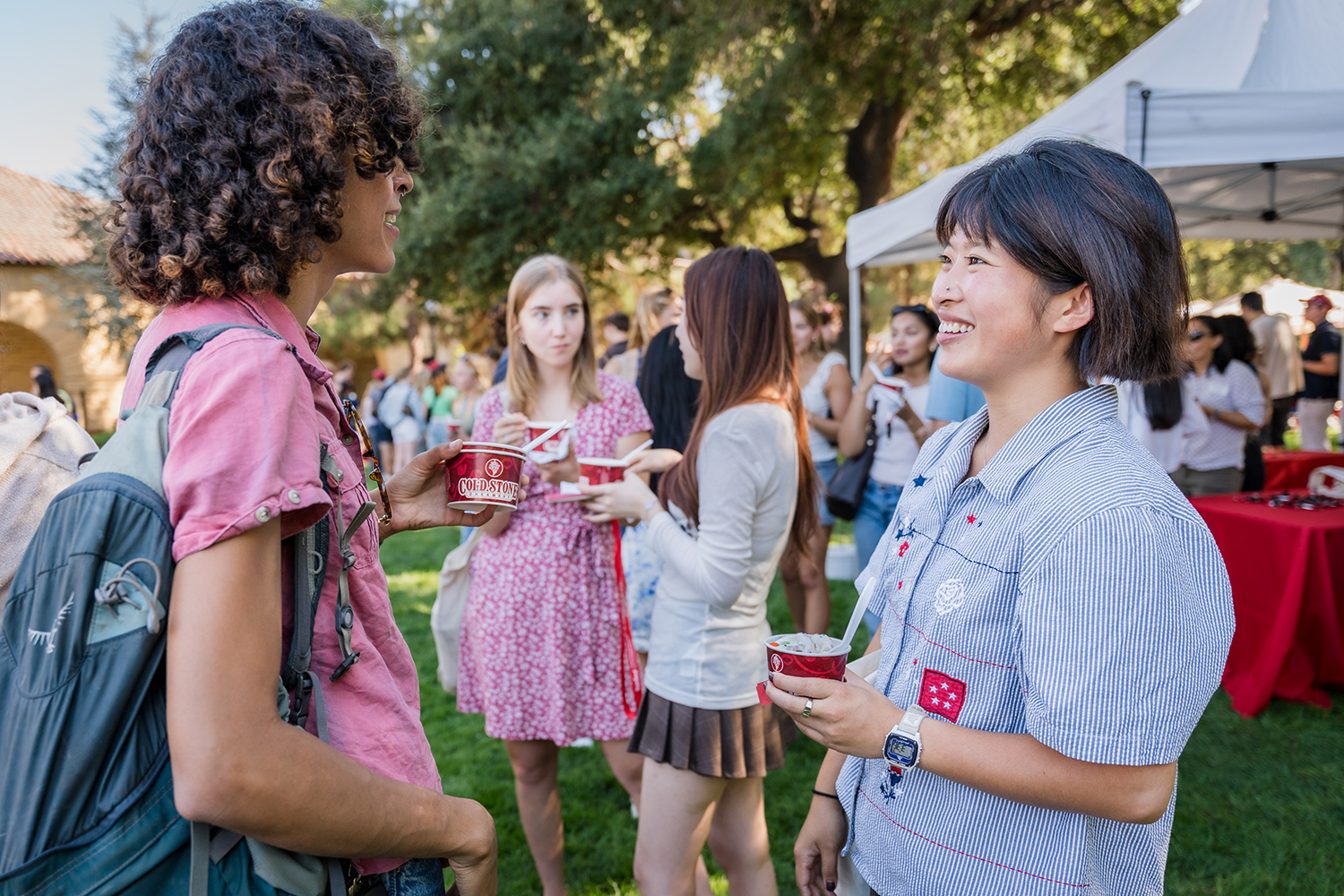 A group of students gather outdoors at an event, enjoying ice cream and socializing under trees and tents.