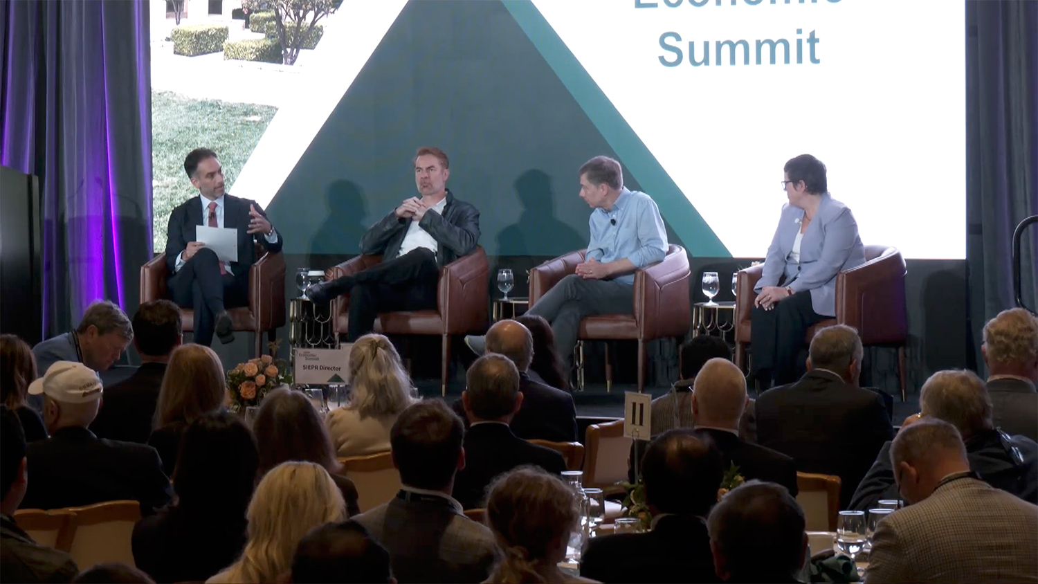 A panel discussion at the 2026 SIEPR Economic Summit, featuring four speakers engaging with an audience seated at round tables.