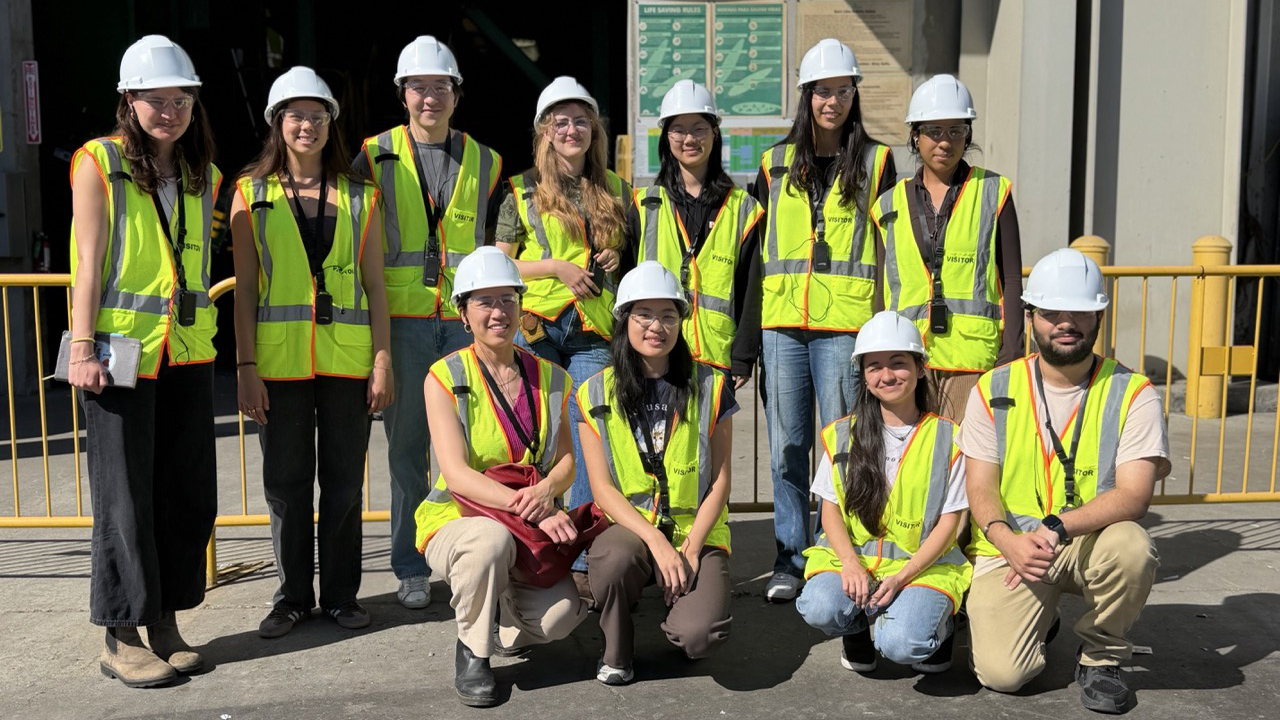Group of eleven people wearing white hard hats and bright yellow safety vests pose together at a construction site.