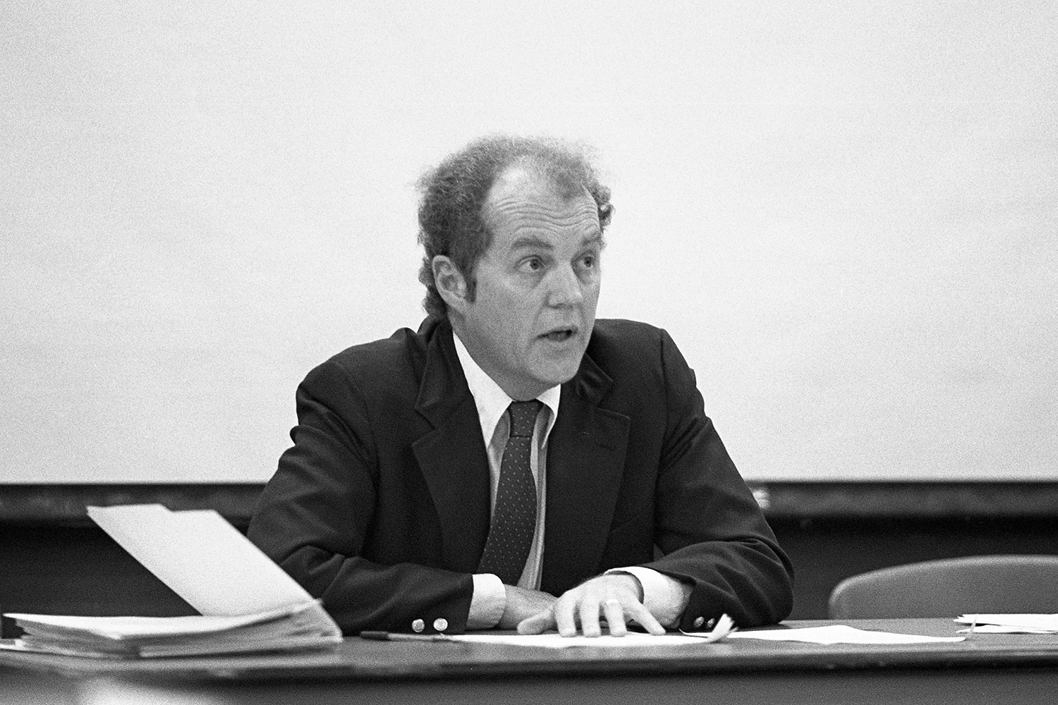 David Abernethy in a suit sitting at a table covered with papers, engaged in discussion, against a plain background.