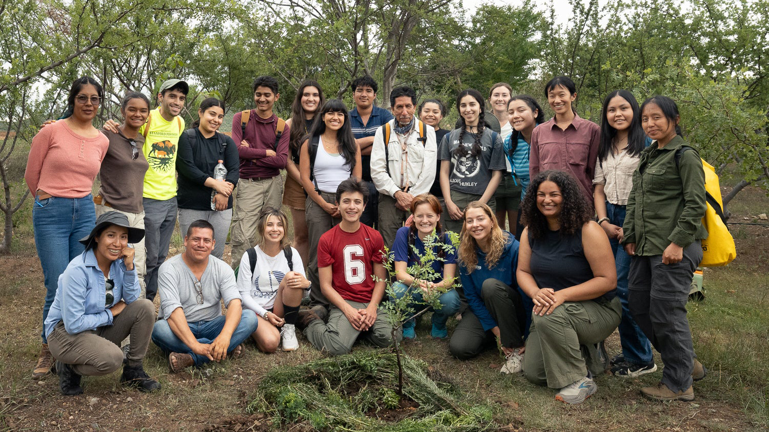 A group of young adults gathers outdoors, posing near a newly planted tree in a natural environment.