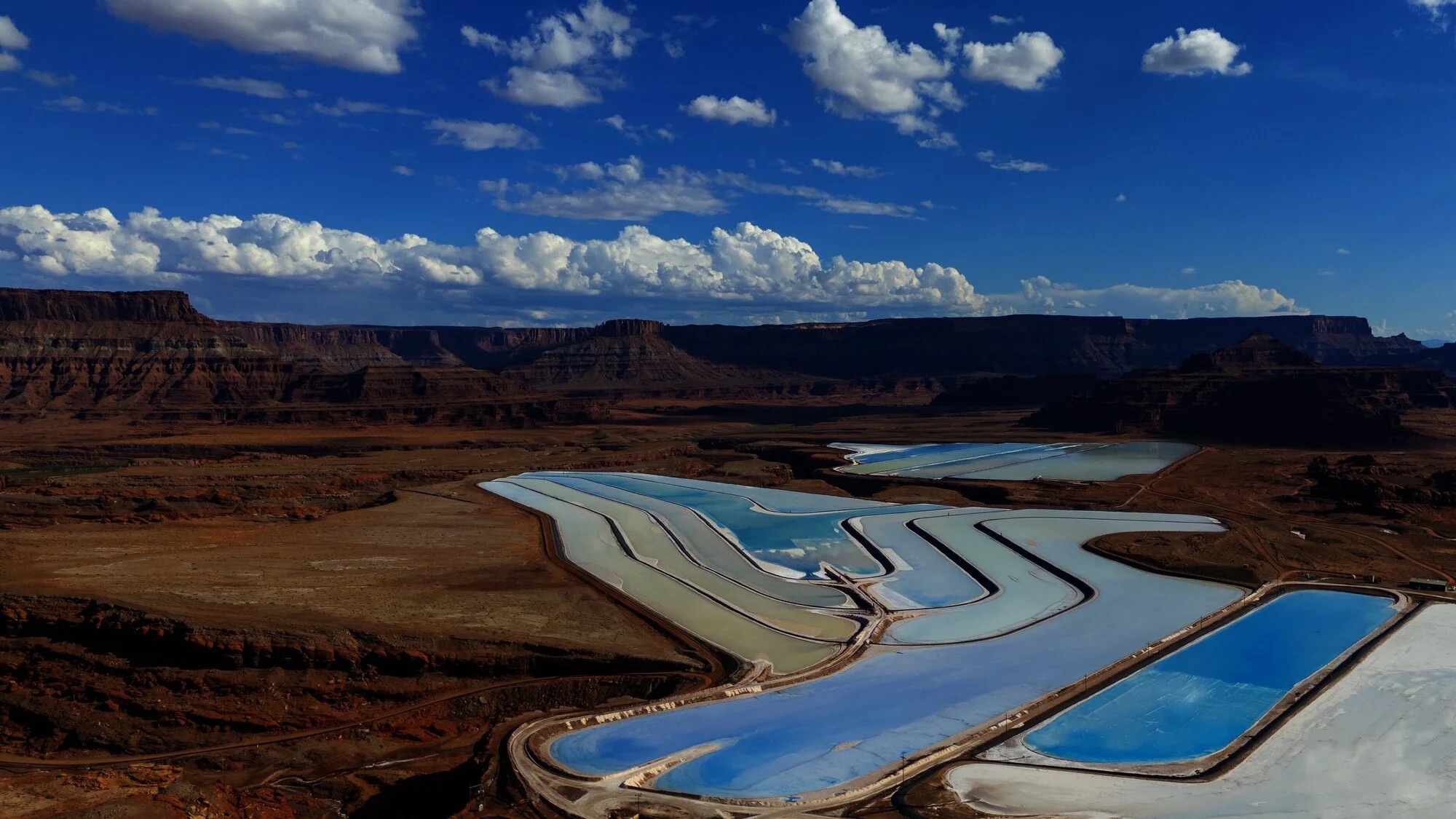 Brightly colored salt evaporation ponds are visible in a rugged landscape, surrounded by cliffs and a clear blue sky with white clouds.