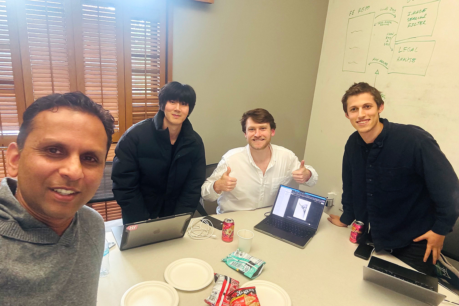 Four young adults pose for a selfie in a meeting room with laptops, snacks, and a whiteboard in the background.
