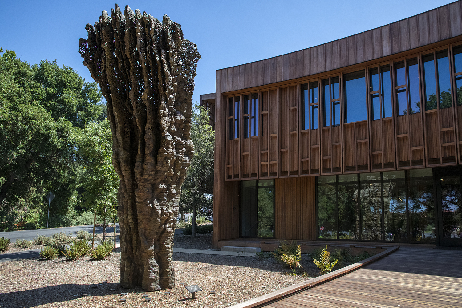 Sculpture resembling a tree stands beside a modern wooden Denning House building amidst landscaped greenery.