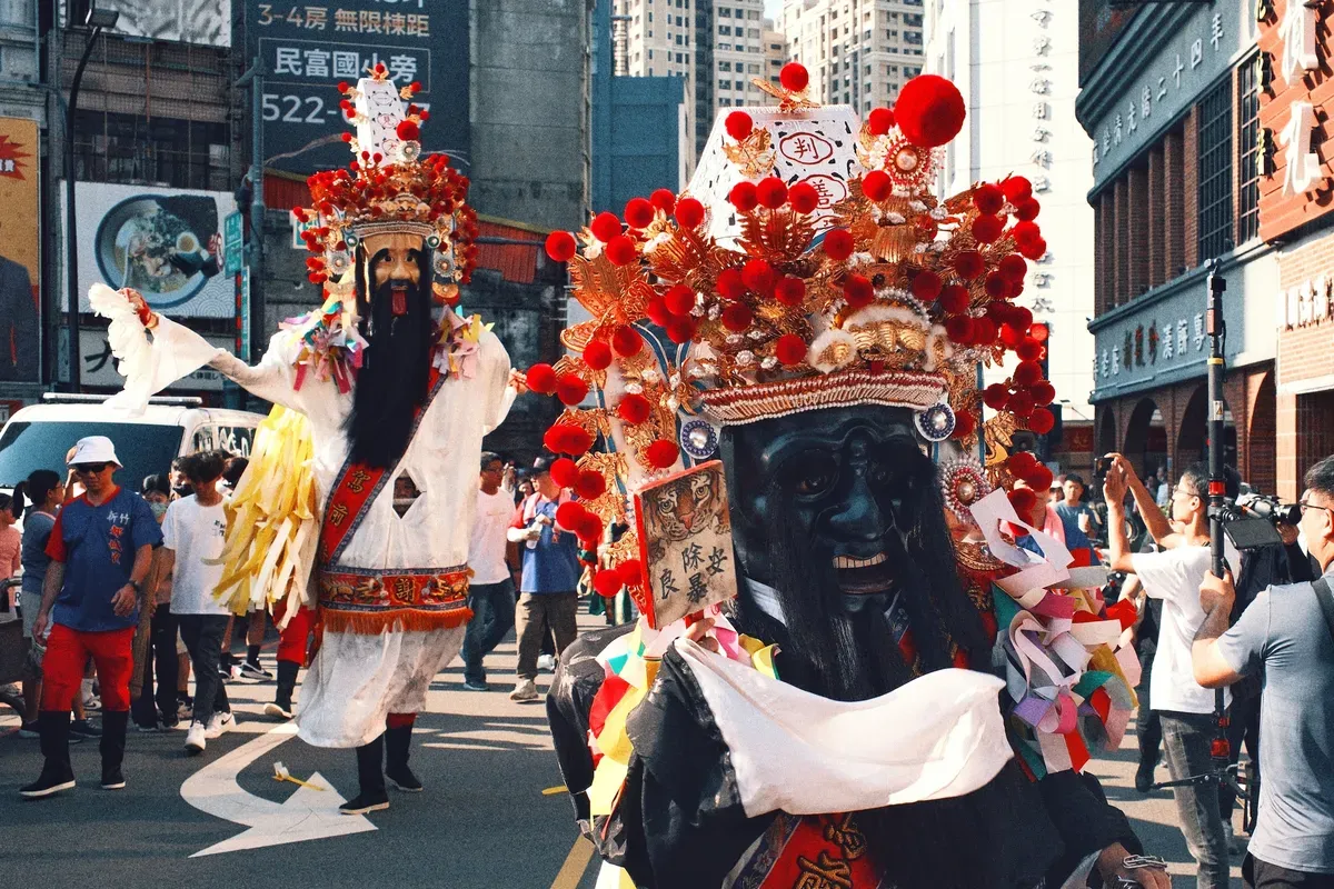 Participants in costumes with colorful headdresses march in a festive street parade surrounded by an audience.