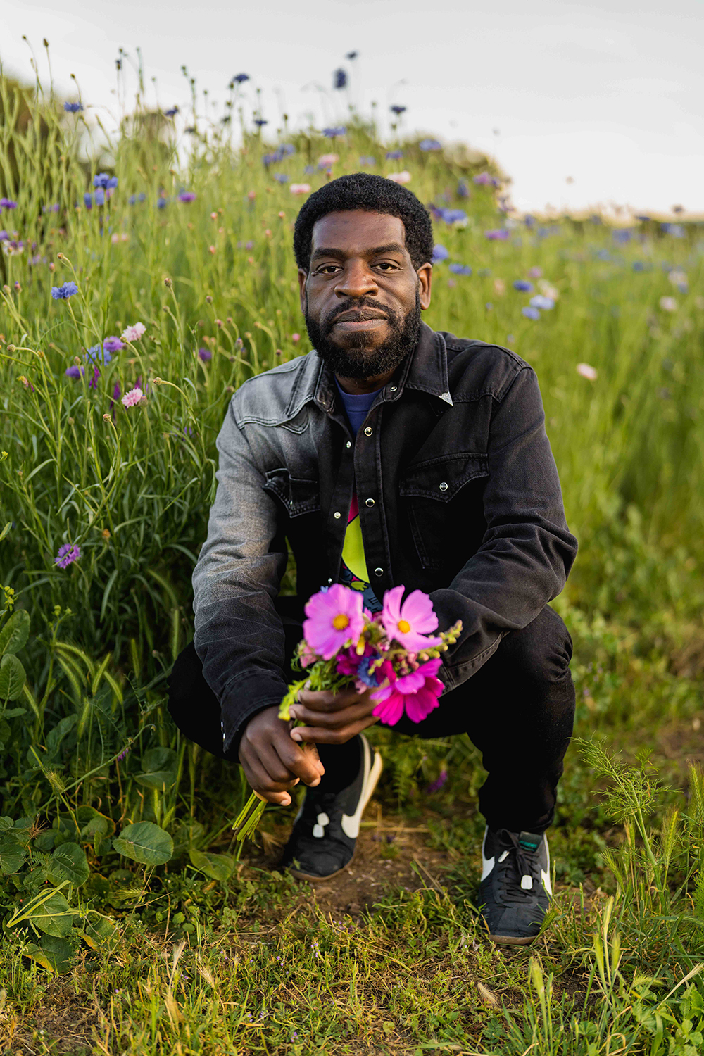 Hanif Abdurraqib crouches in a field of flowers, holding a bouquet of colorful blooms and looking at the camera.
