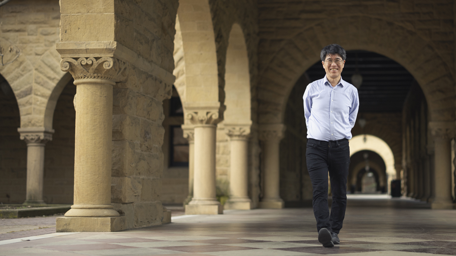 Tadashi Tokieda walks through an archway in a stone courtyard, surrounded by columns and historic architecture.