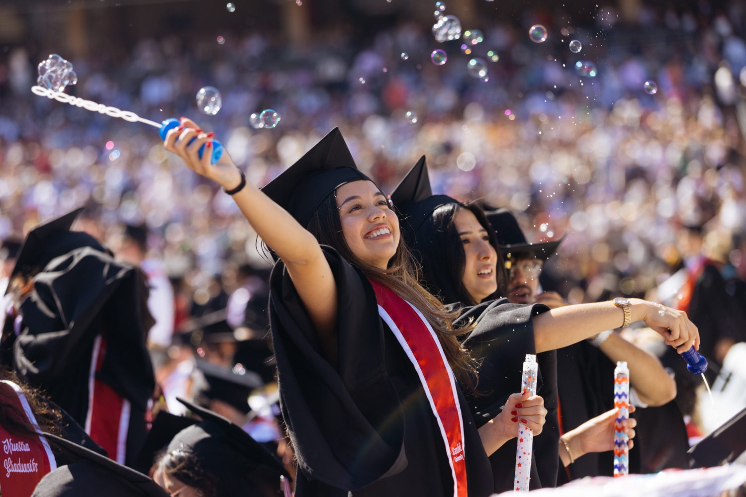 Graduate in cap and gown blows bubbles during commencement ceremony as crowd celebrates in background.