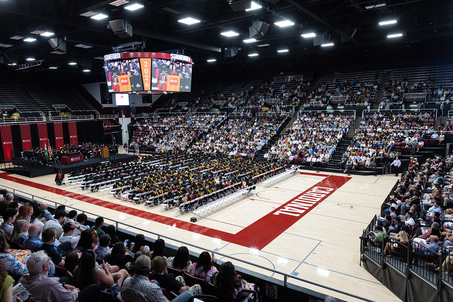 Graduates in caps and gowns seated on basketball court floor with audience members filling the surrounding stands.