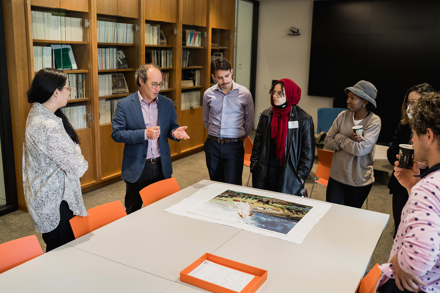 Linetzky and a group of students discuss a print displayed on a table in a meeting room, with bookshelves visible in the background.