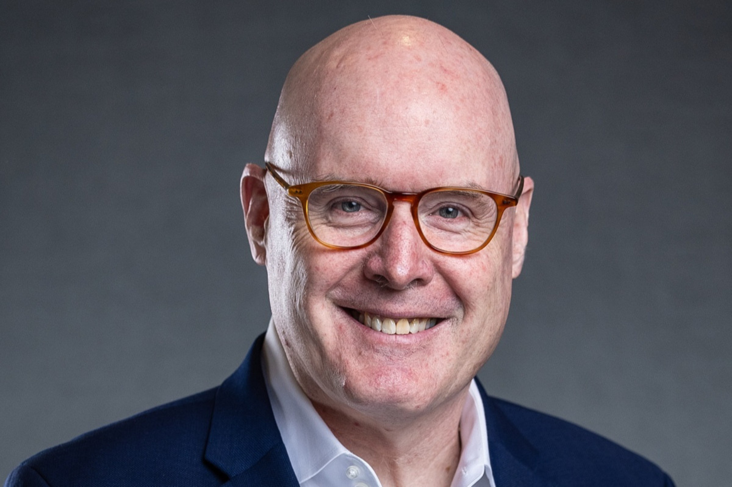 Headshot of a smiling Timothy Lynch wearing amber-framed glasses and a navy blazer against a gray background.