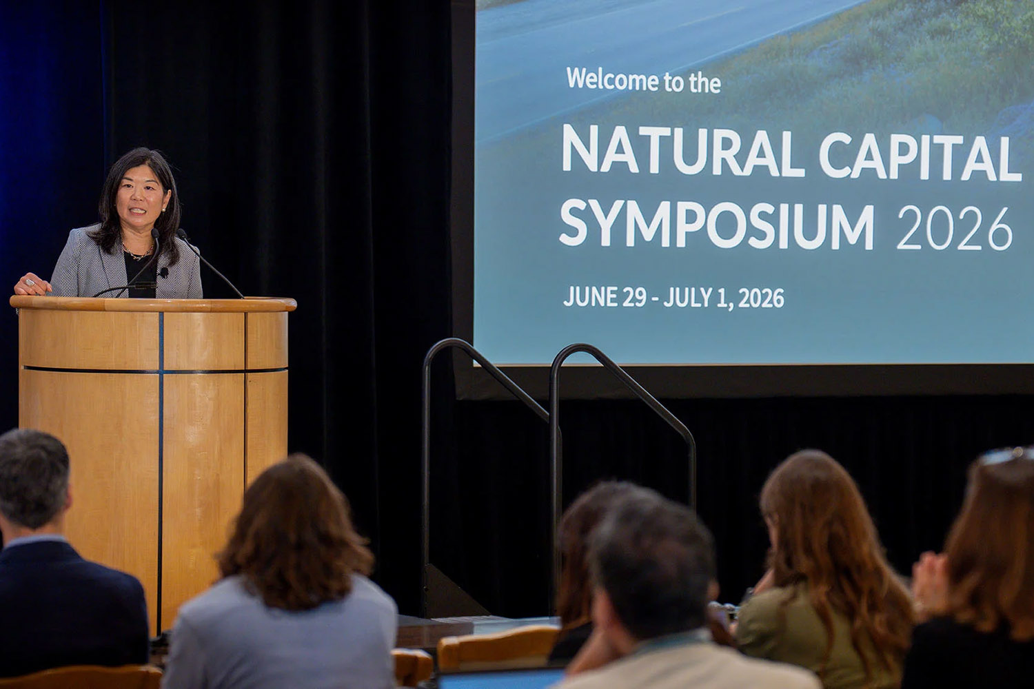 A woman speaks at a podium during the Natural Capital Symposium 2026, with a large welcome banner displayed behind her.