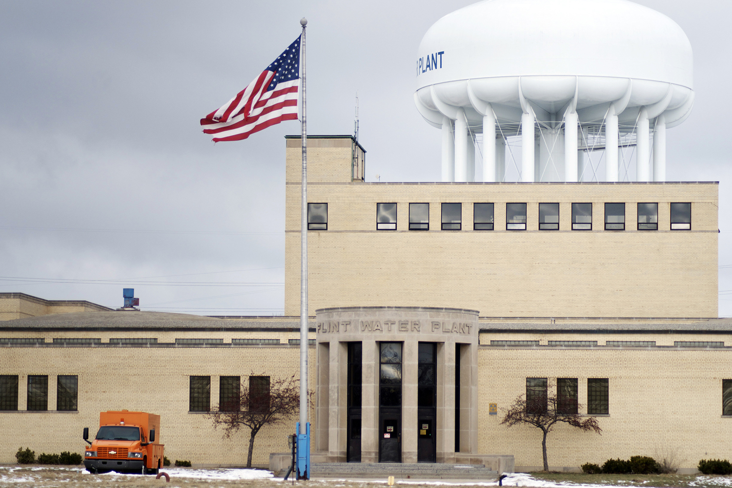 The Flint Water Plant, a tan brick building with a large white water tower behind it and a U.S. flag flying out front under an overcast sky.