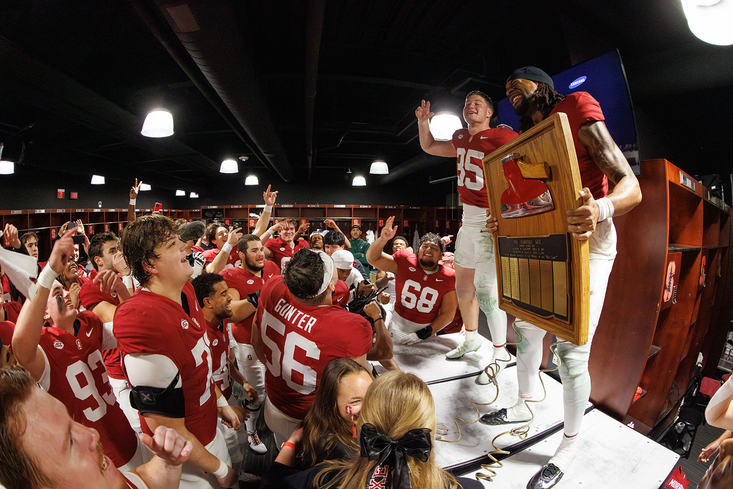 Cardinal football players celebrate in the locker room, holding up a wooden axe trophy after a win.