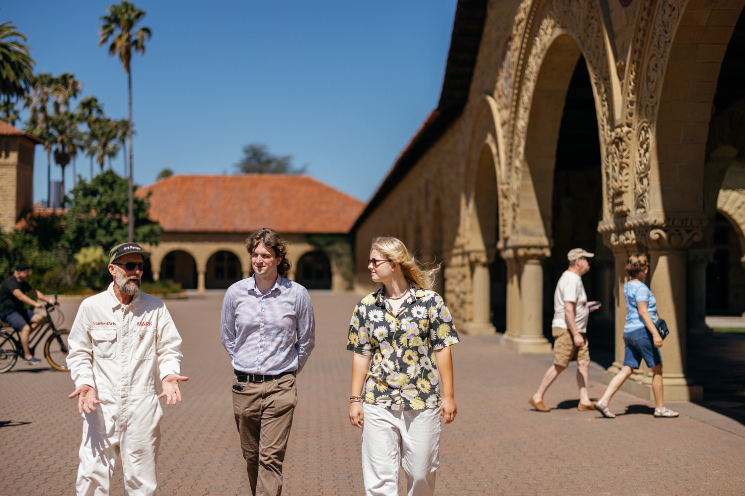 Mark Shunney in white coveralls talks with two visitors while walking past sandstone arches on a sunny Main Quad.