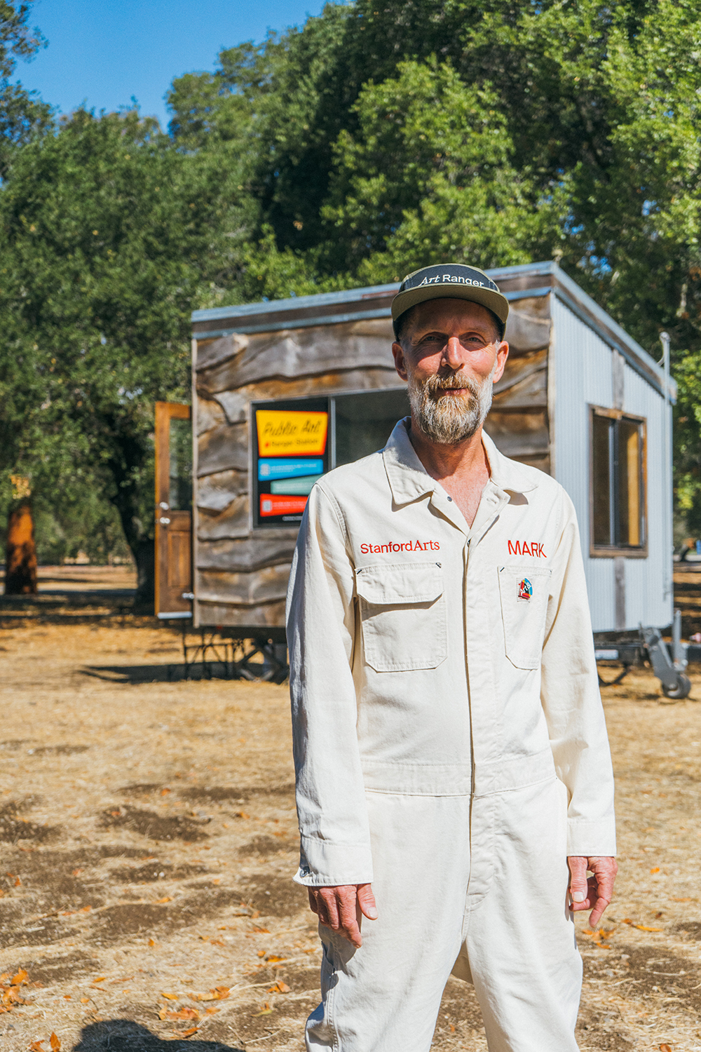 Mark Shunney in a Stanford Arts coverall stands outside a small trailer marked as a public art ranger station, under oak trees.