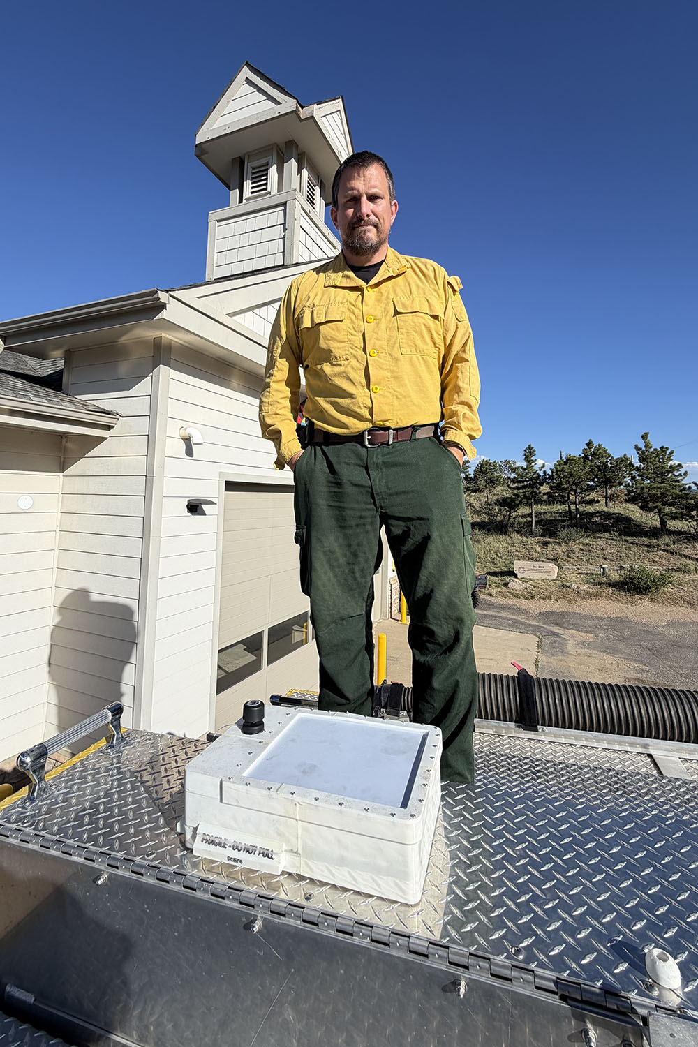Firefighter in yellow shirt and green pants stands atop a fire truck beside a white sensor unit.