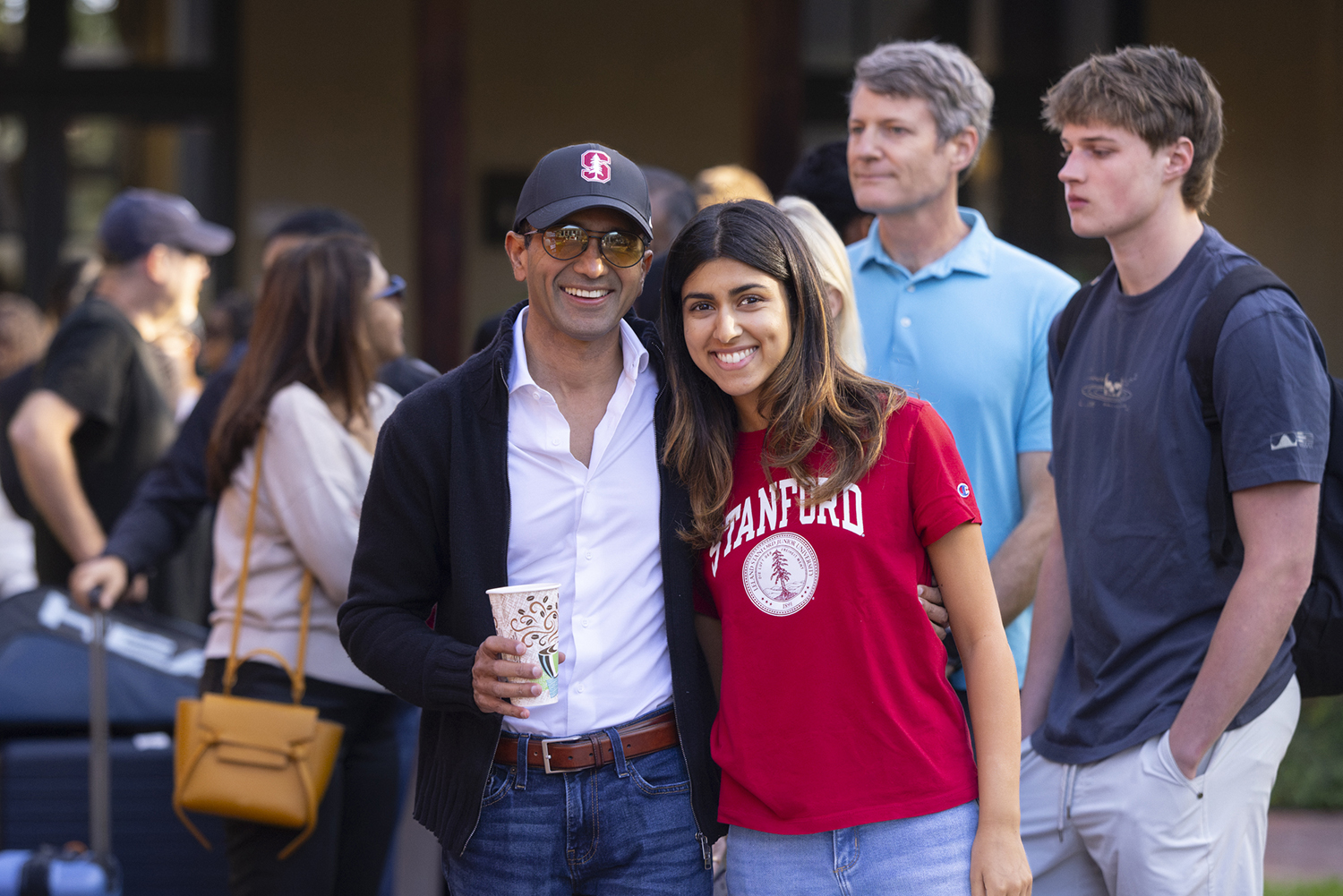 A father and his student pose together outside a residence hall as other families gather in the background.
