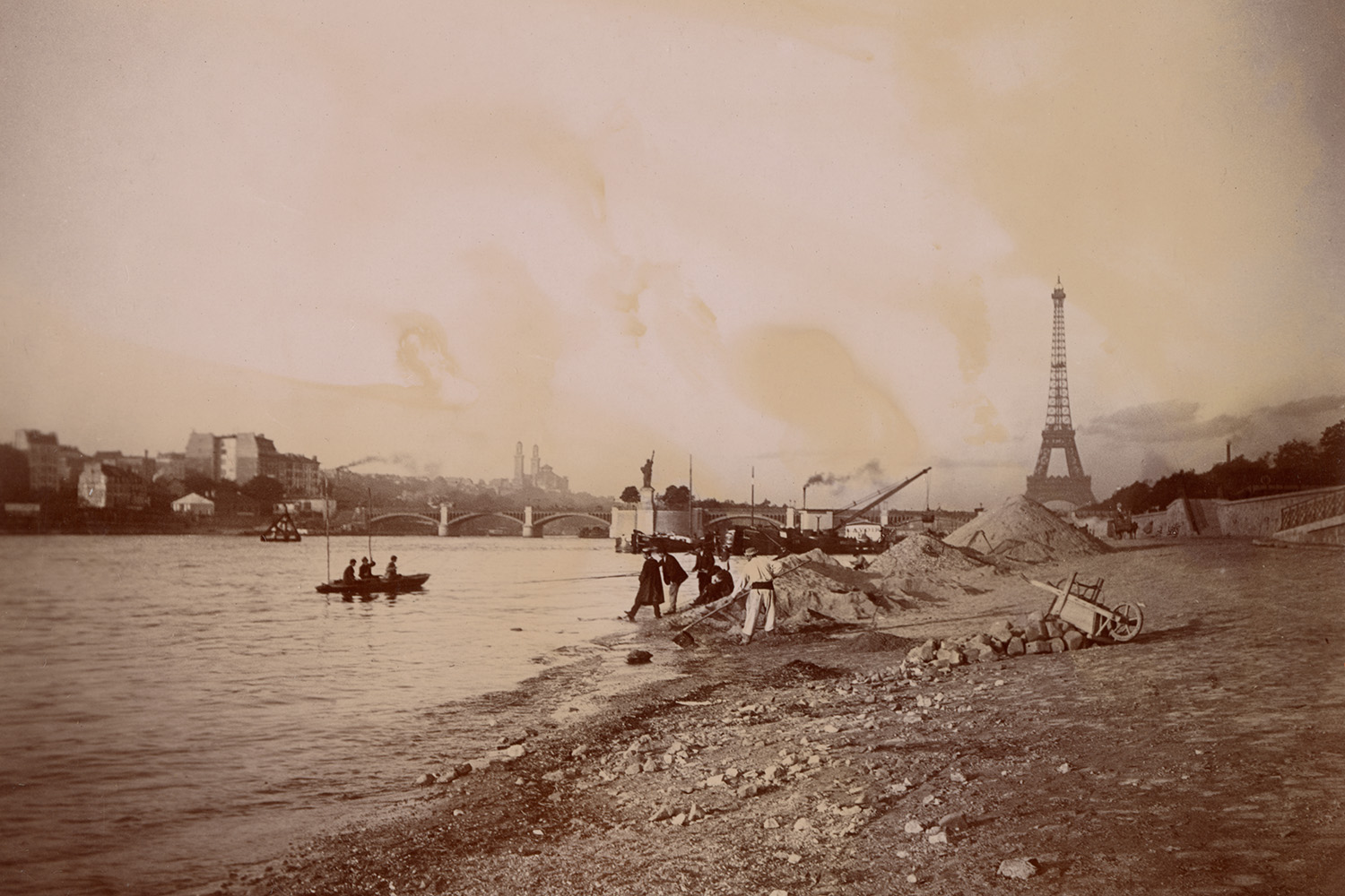 A vintage photograph shows the Eiffel Tower under construction beyond the Seine's rocky riverbank.