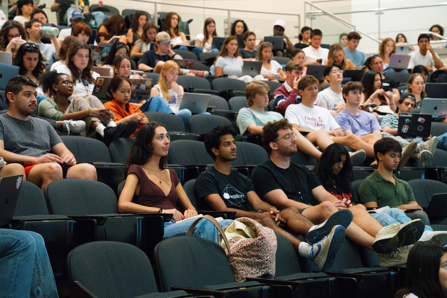Students fill the tiered seats of a lecture hall, many with laptops open, listening to a presentation off camera.