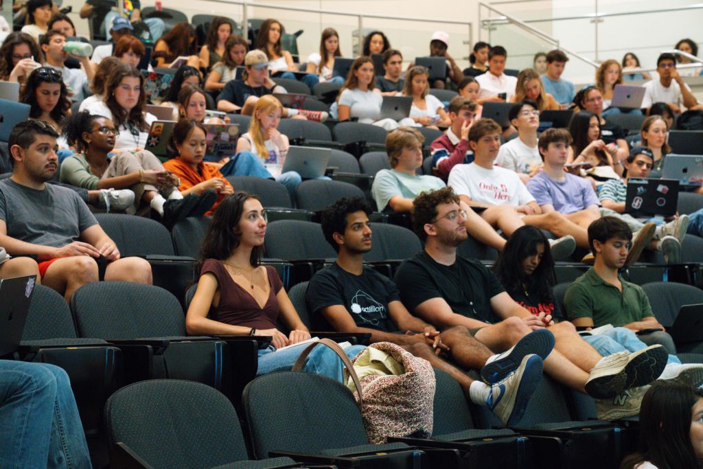 Students fill the tiered seats of a lecture hall, many with laptops open, listening to a presentation off camera.