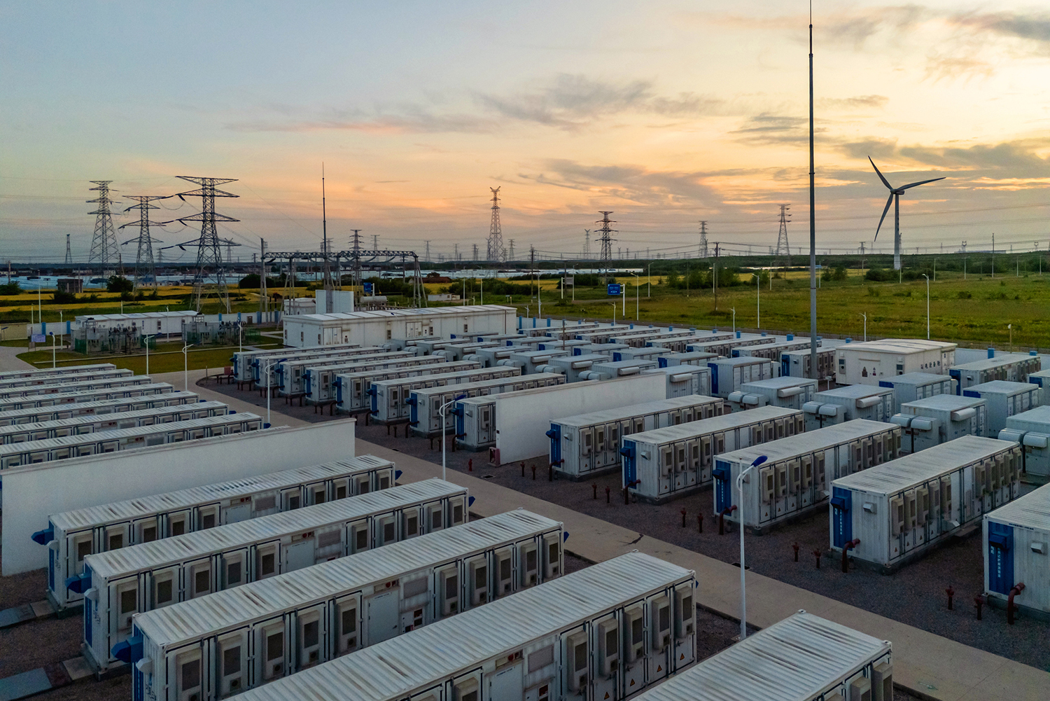 Rows of white container-sized units at an energy storage facility, with power lines and a wind turbine in the distance at sunset.