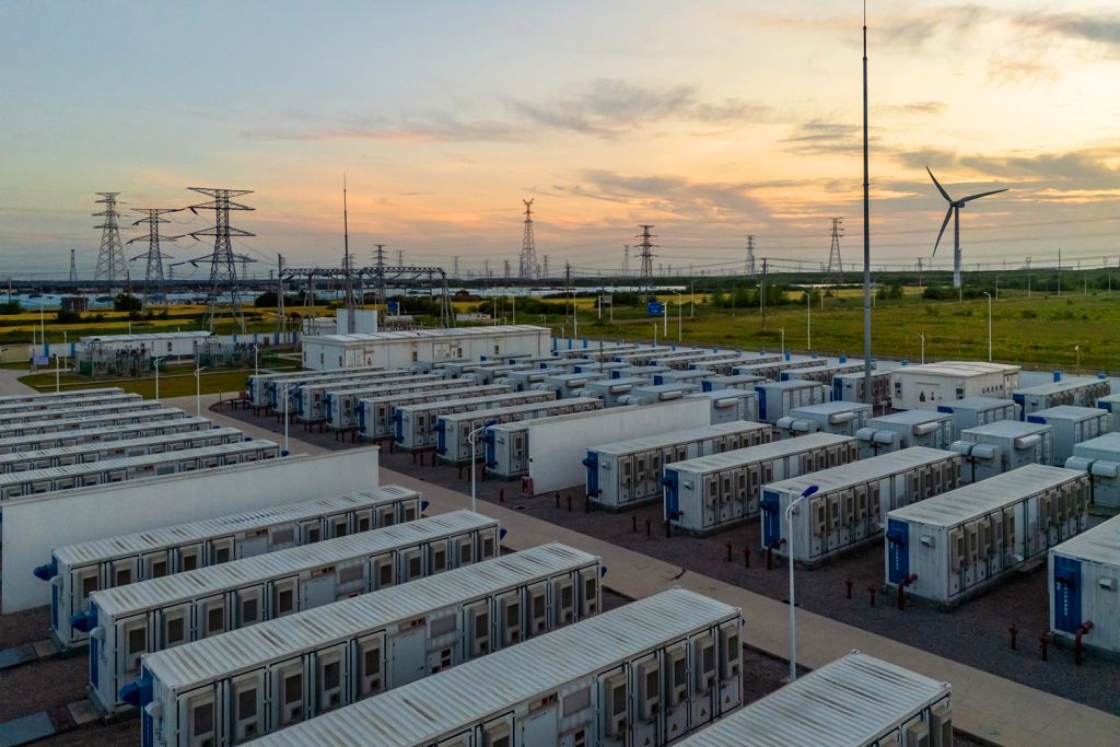 Rows of white container-sized units at an energy storage facility, with power lines and a wind turbine in the distance at sunset.