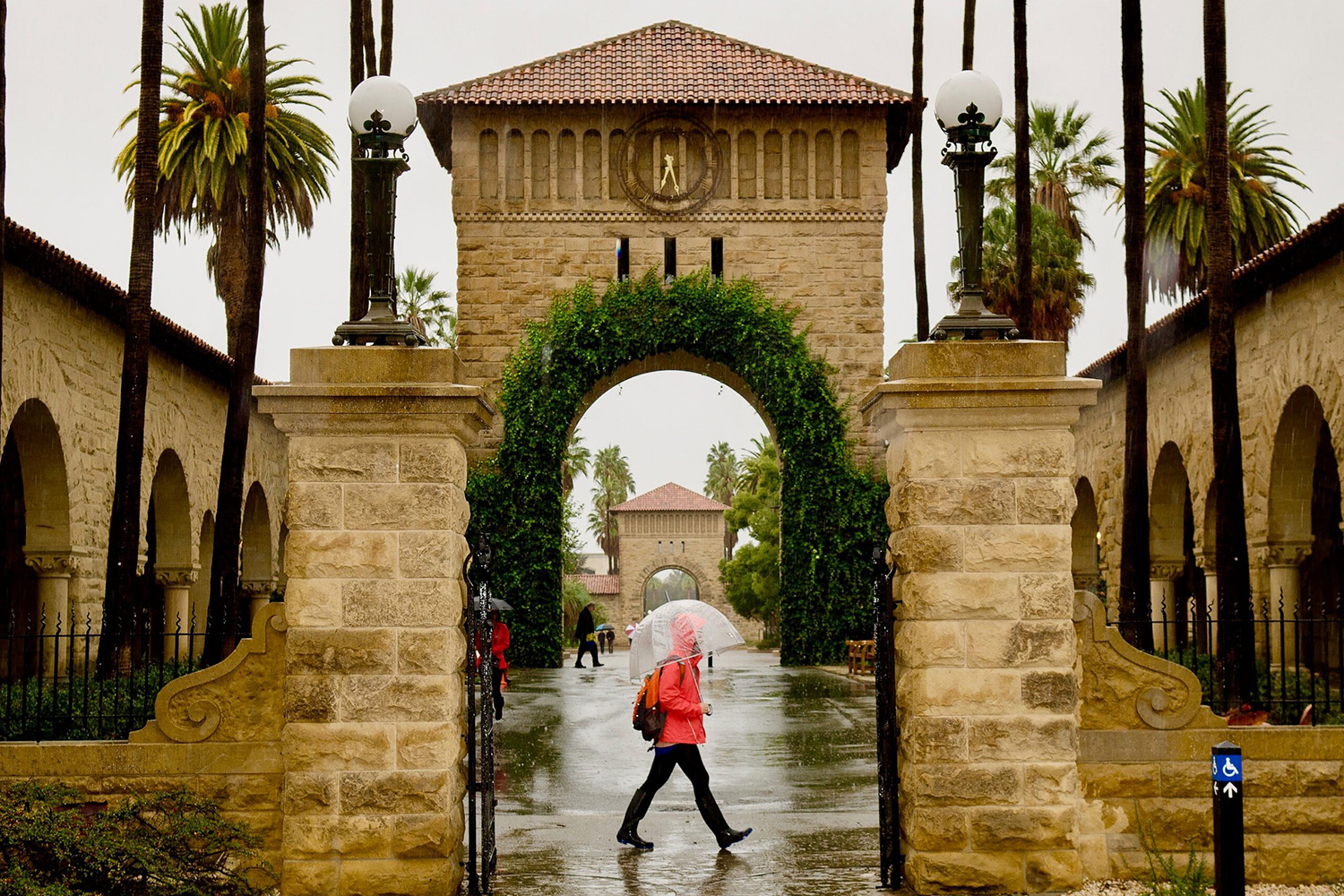 A student with a clear umbrella walks through a rainy Stanford Main Quad gate beneath an ivy-covered sandstone arch.