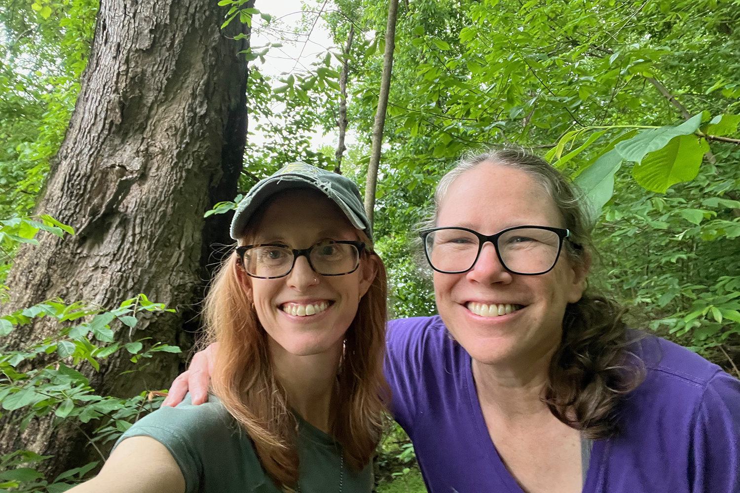 Two smiling women pose close together for a selfie in a green forest, one in a cap and one in a purple shirt.