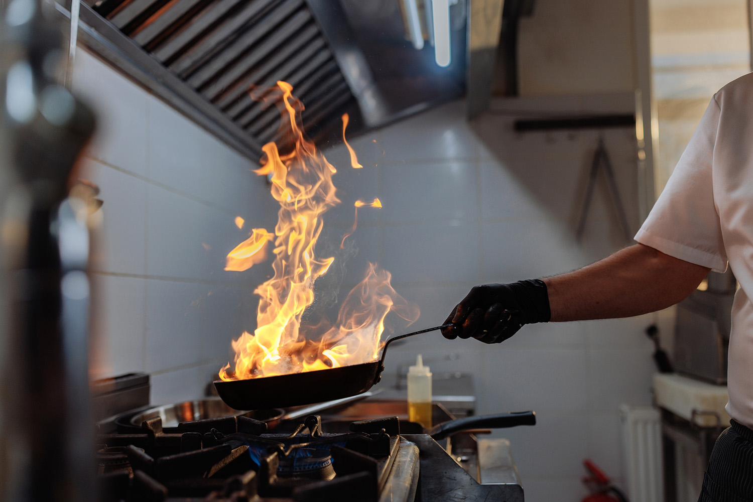 A gloved chef holds a flaming pan over a gas stove's blue flame in a restaurant kitchen.