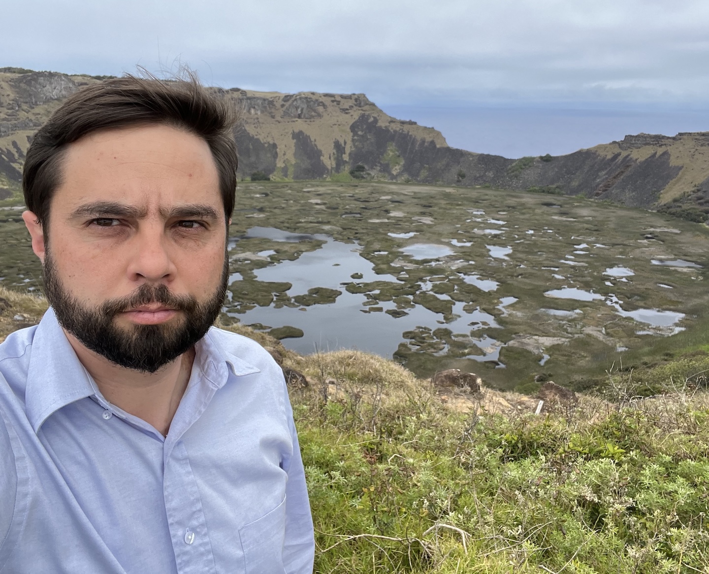 Alexander Ioannidis takes a selfie on a grassy rim above a marshy crater lake with the ocean beyond.
