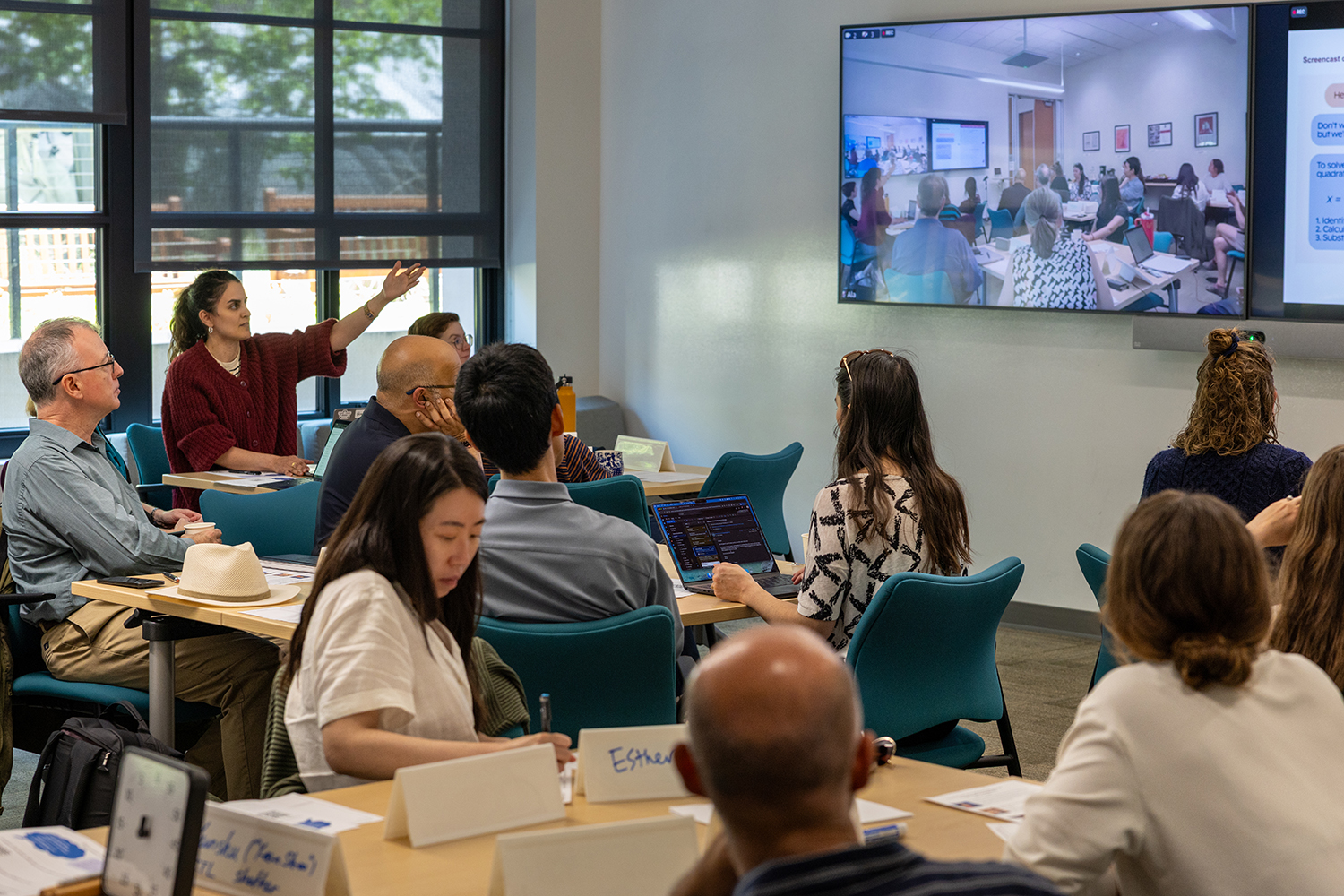 Shima Salehi points to a screen showing a video call with another classroom as seated participants watch.
