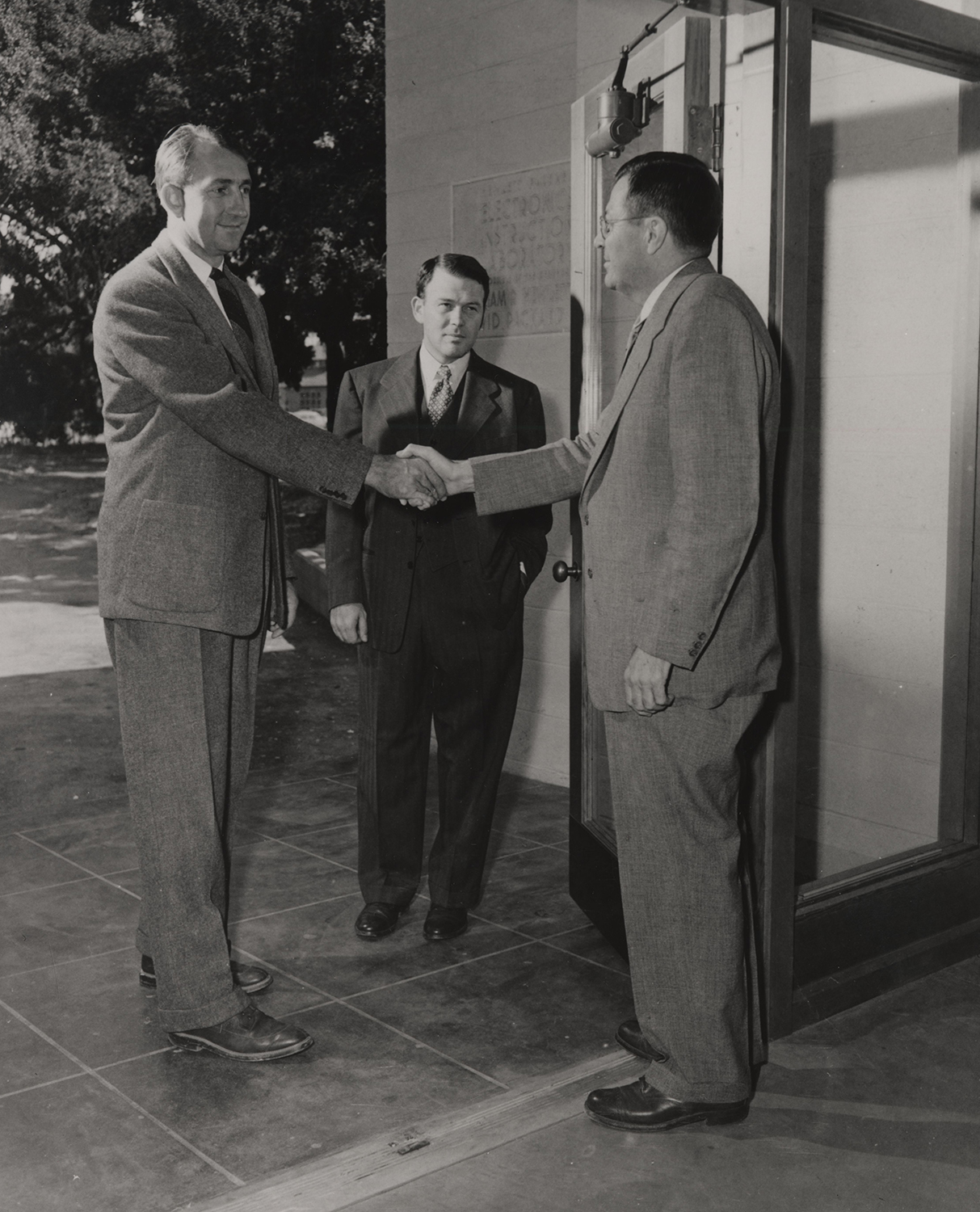 David Packard and Fred Terman shake hands with Bill Hewlett looking on at entrance to a laboratory building.