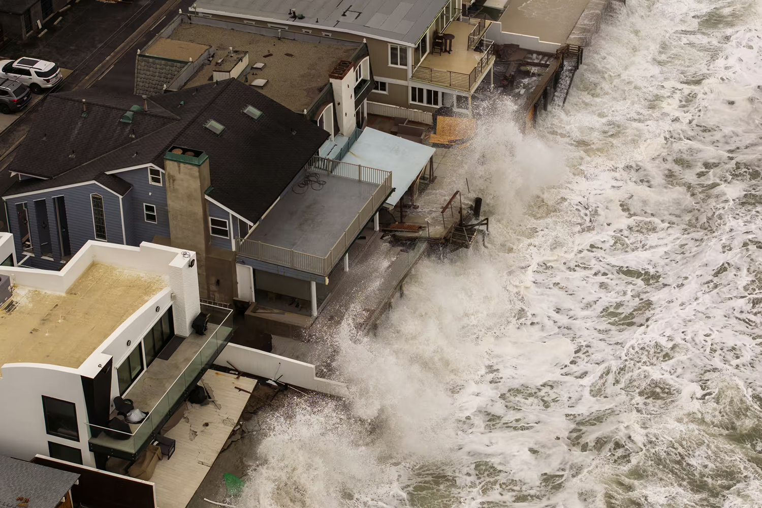 Aerial view of large waves battering oceanfront homes, with storm damage visible on decks and walkways.