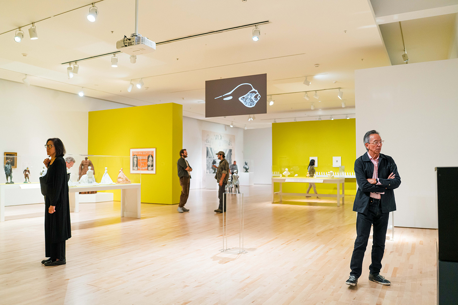 Museum visitors walk through a gallery with yellow accent walls, display cases of small sculptures, and framed prints.