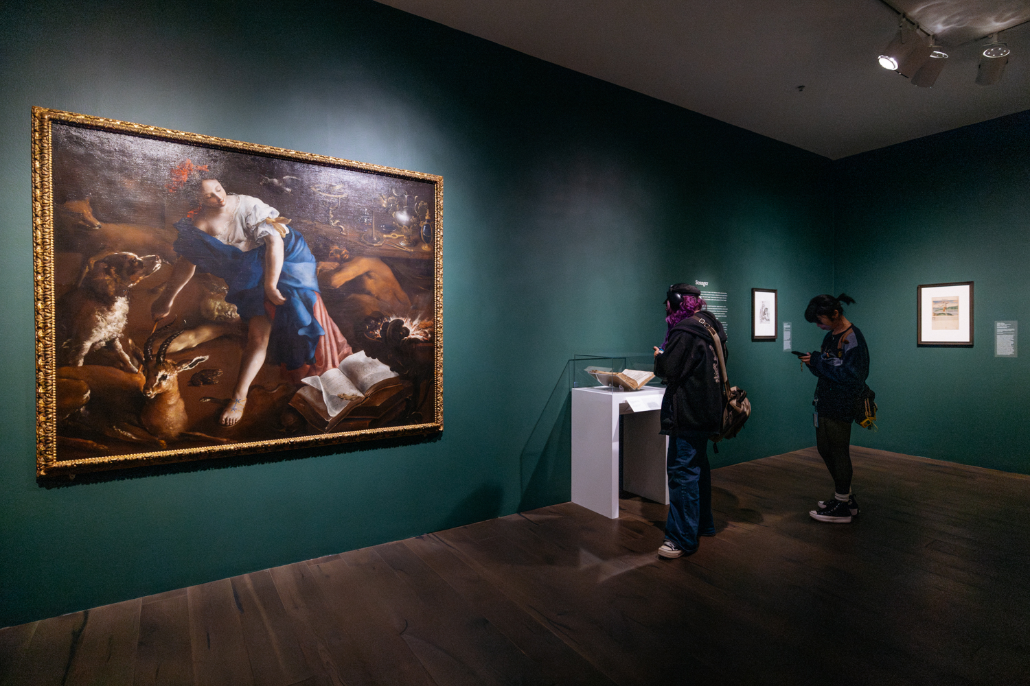 The 17th-century oil painting 'The Sorceress' by Bartolomeo Guidobono, depicting a woman surrounded by animals and magical elements, is displayed on a dark green museum gallery wall. Visitors observe nearby objects in a glass case.