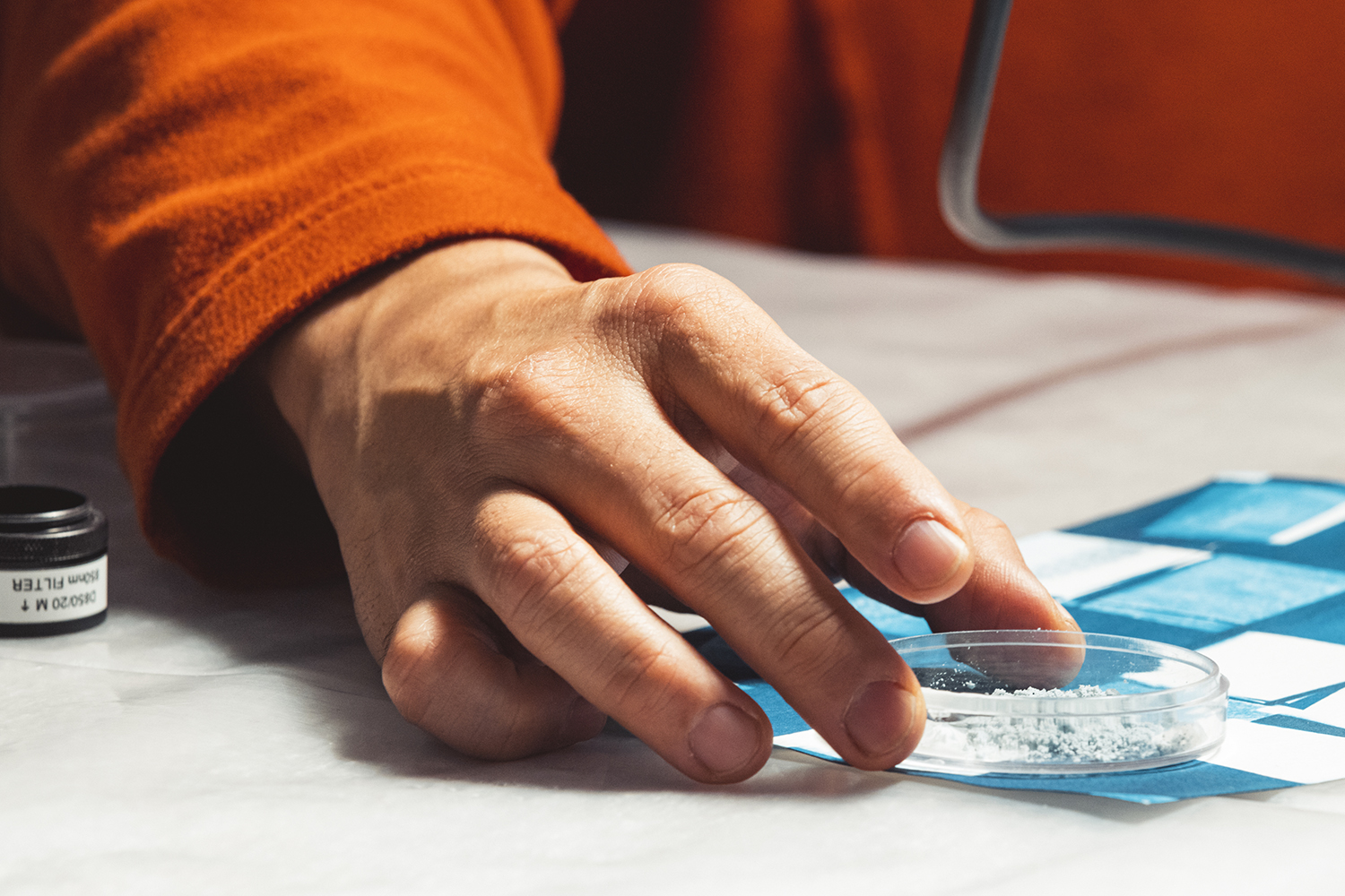 A hand carefully places a petri dish with white powder on a blue grid, indicating a scientific procedure or experiment.