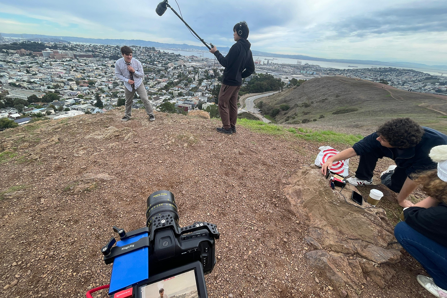 A group films outdoors on a hilltop, with a scenic view of the city and a person preparing for a scene and another person holding a boom mic.