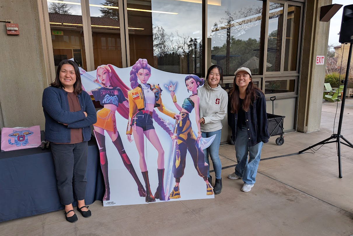 Four individuals pose next to a large cutout of colorful animated characters at an event featuring K-Pop.