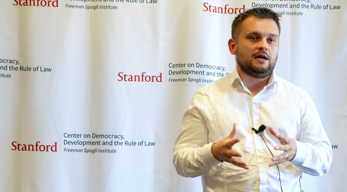Mykhailo Pavliuk gestures while speaking at a podium in front of a backdrop featuring logos of the Stanford Center on Democracy and the Rule of Law.