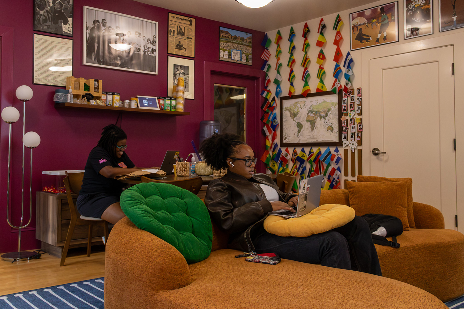 A cozy room featuring colorful wall art, flags, and two people working on laptops from comfortable seating.
