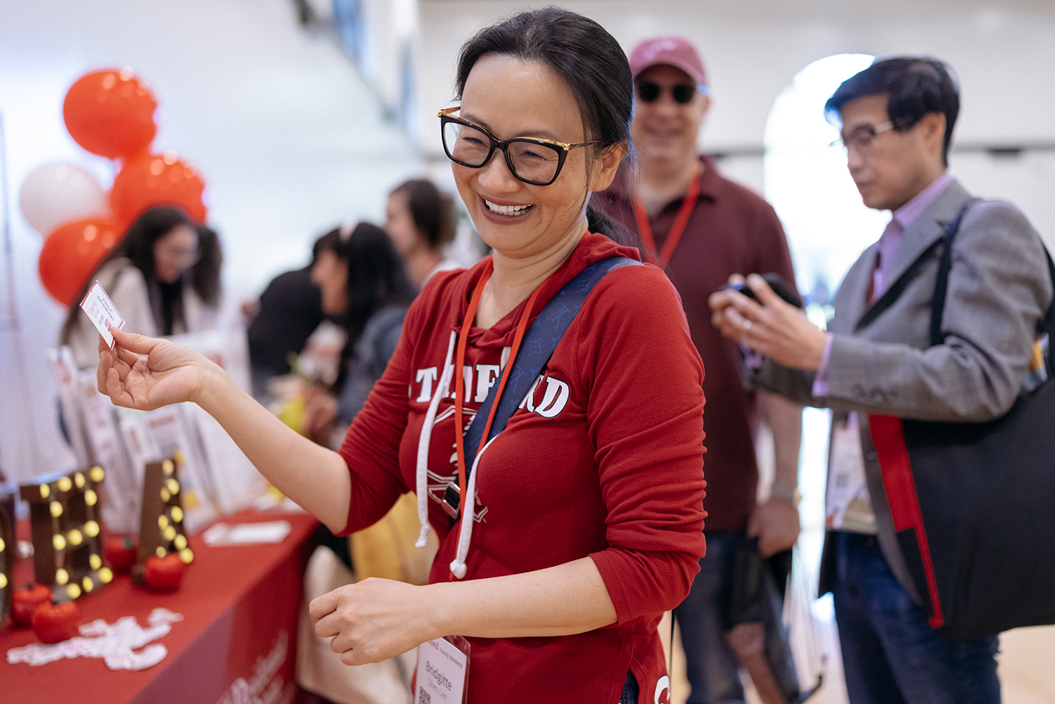 A woman in a red Stanford hoodie holds a card while engaging with attendees at a booth decorated with balloons and promotional materials.