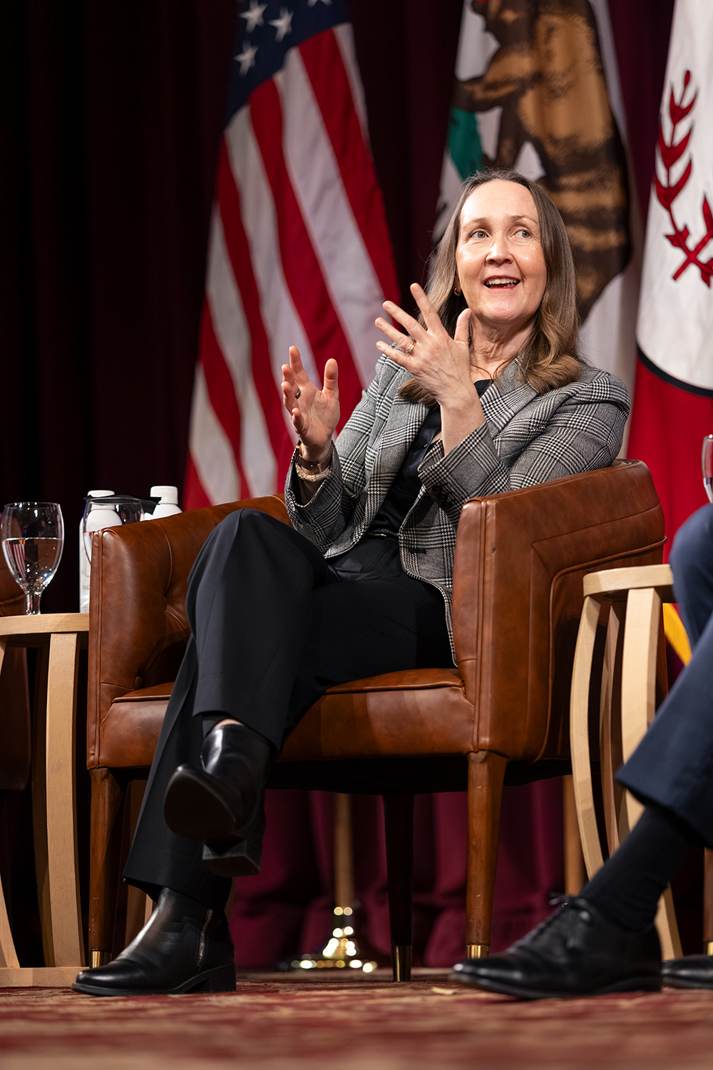 Jenny Martinez gestures during a panel discussion, seated in an armchair with water and microphones on a table nearby, flanked by flags.
