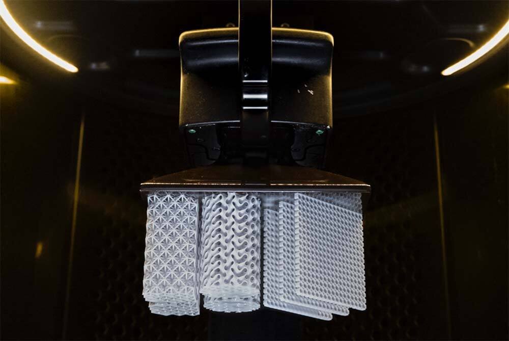 Close-up of a 3D printer nozzle with three transparent, patterned test prints suspended below it.