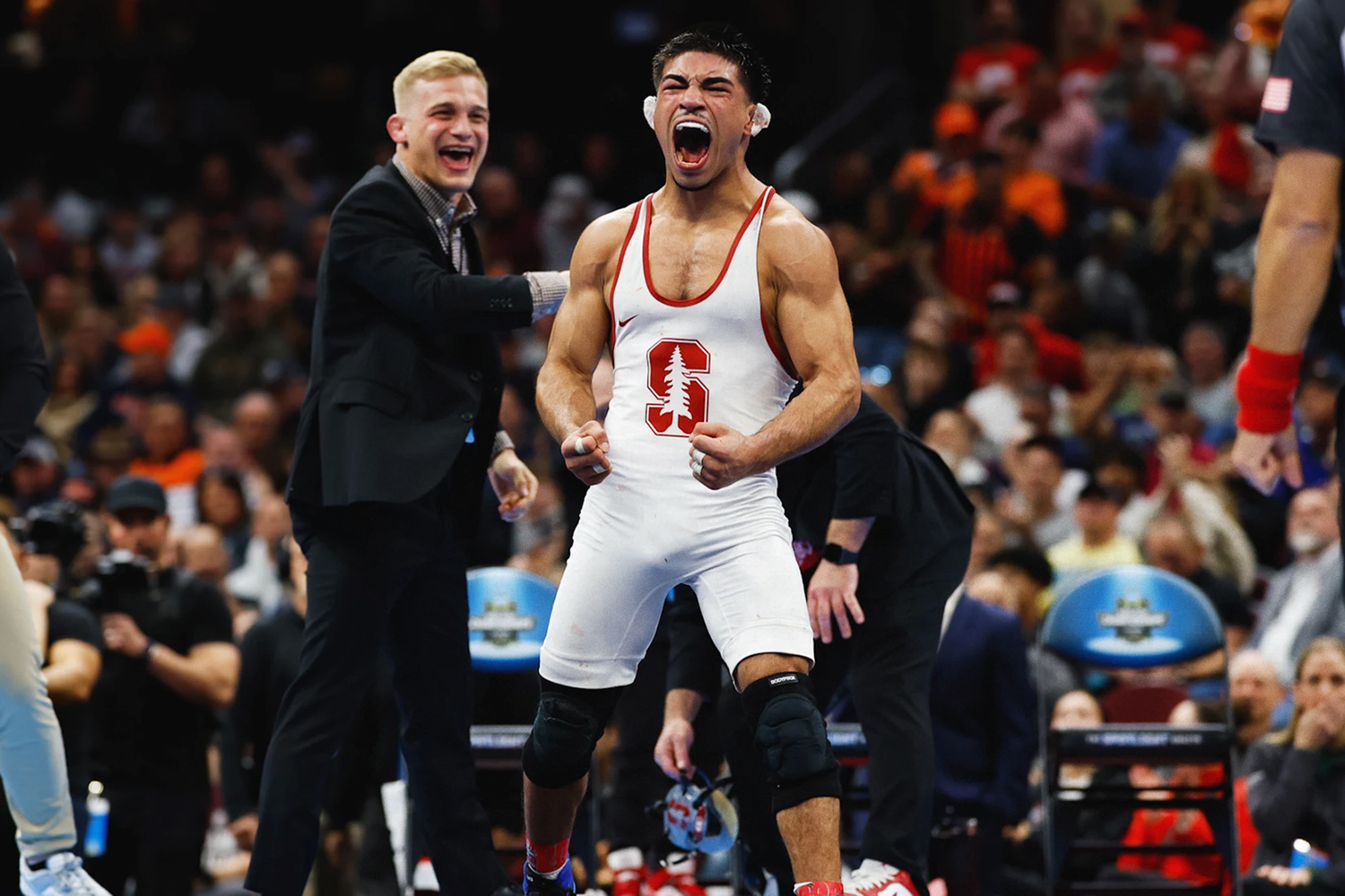 Aden Valencia in a red and white singlet celebrates in a competitive arena, with spectators cheering in the background.