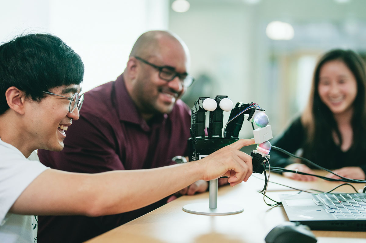 Three people smile while examining a small handlike robotics project on a desk.