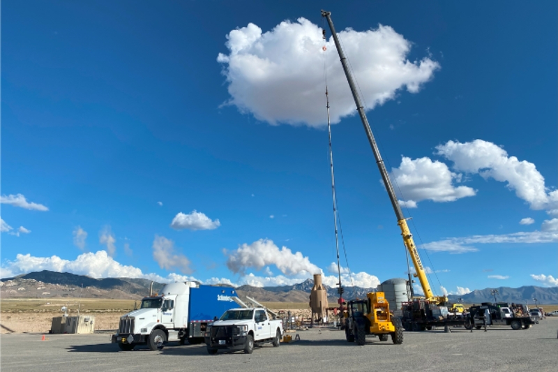Heavy machinery and trucks are present at a work site, with a crane lifting equipment against a clear blue sky and mountains in the background.