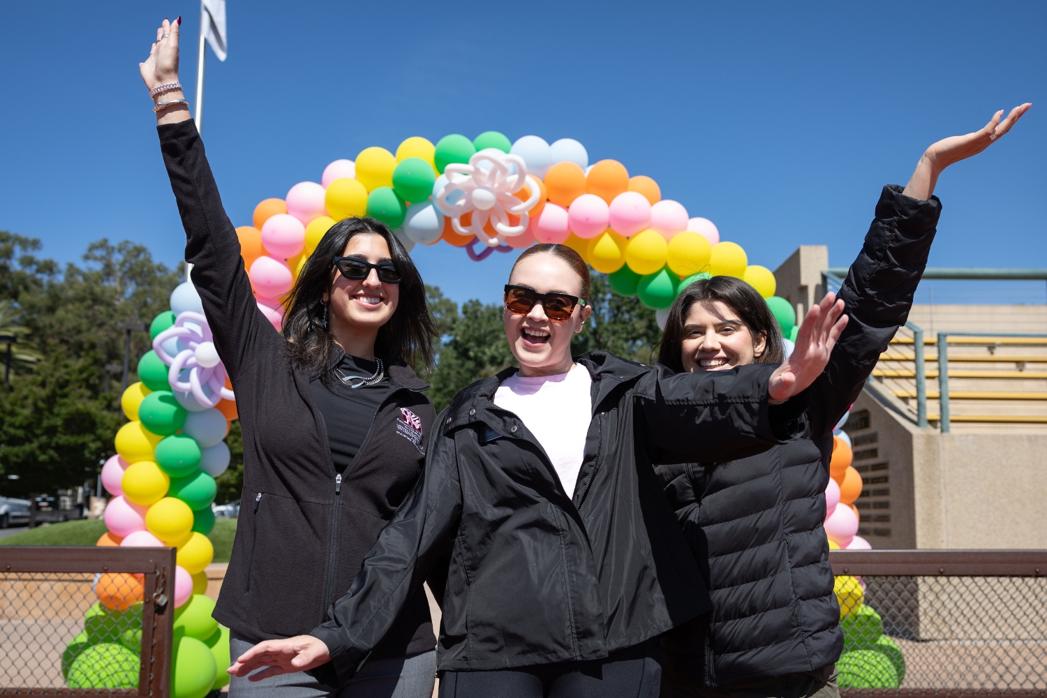 Three people pose joyfully in front of a colorful balloon arch, celebrating a festive event outdoors.
