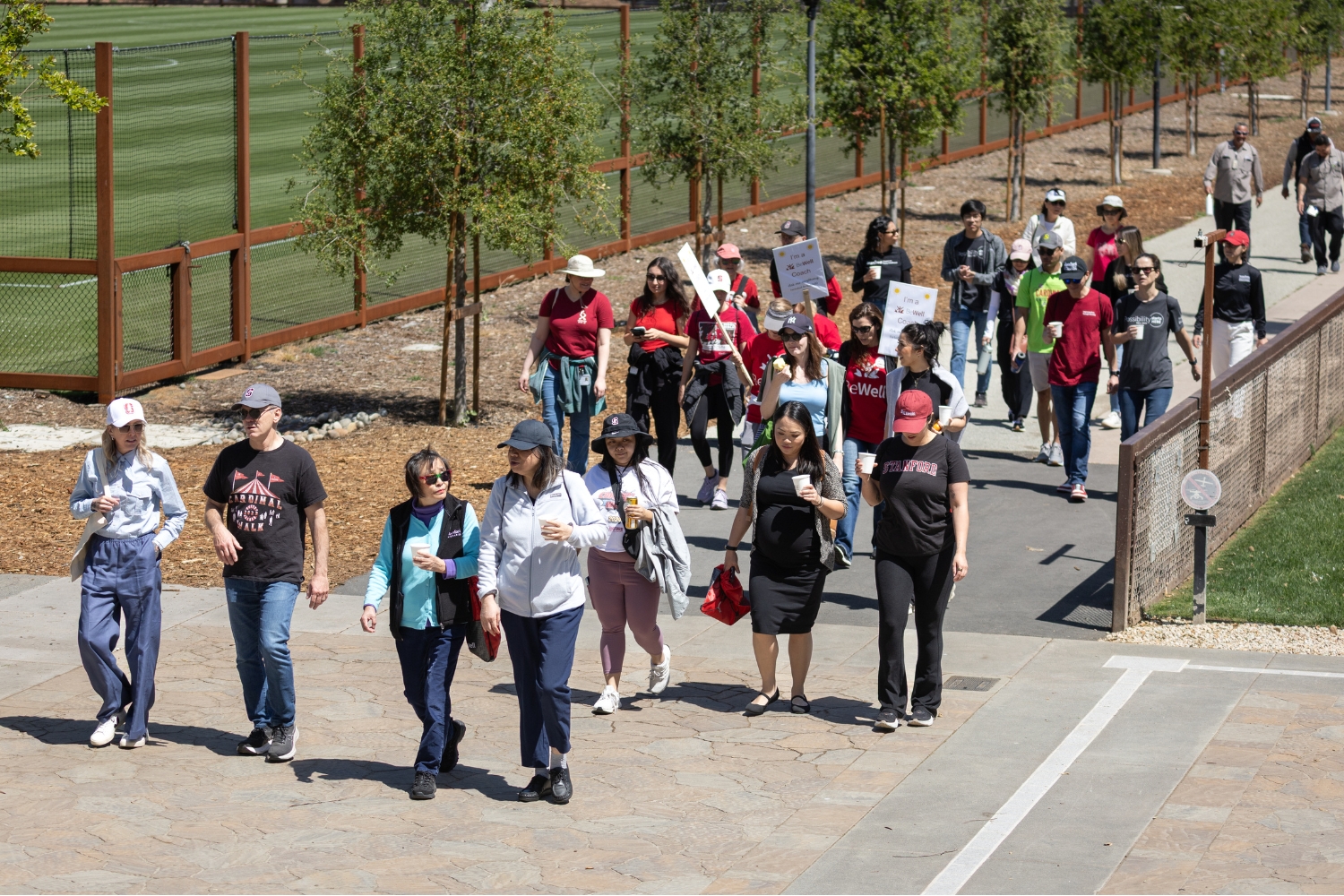 A crowd of people walking along a pathway, holding cups and signs, participating in a community wellness event on a sunny day.